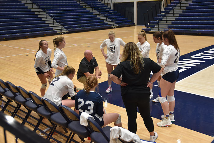 Eight female  volleyball players in a circle with male coach speaking with them while female coach looks on