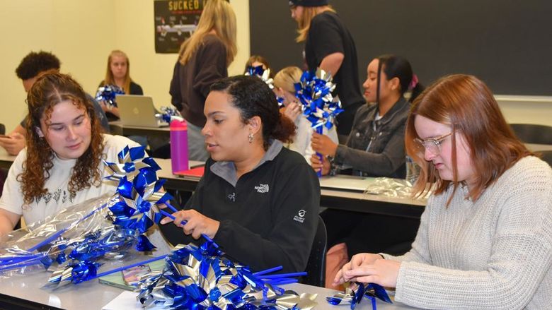 Three females college student at a table assembling pinwheels, male and female classmates in the background doing the same