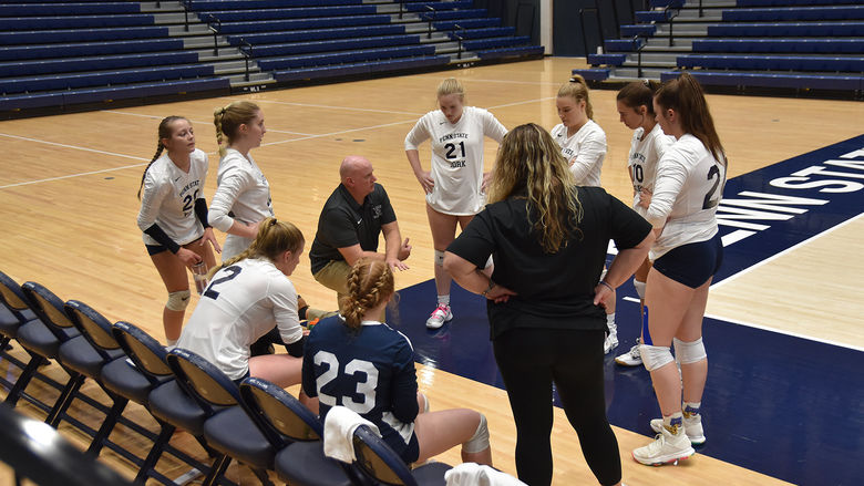 Eight female  volleyball players in a circle with male coach speaking with them while female coach looks on