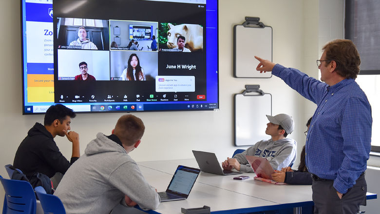 Faculty member pointing at screen with students seated at a table with laptops and others pictured on screen remotely