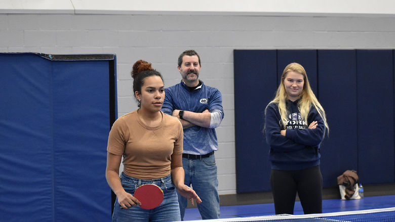 A Latino female college student plays ping pong while an older male and female college student watch. 