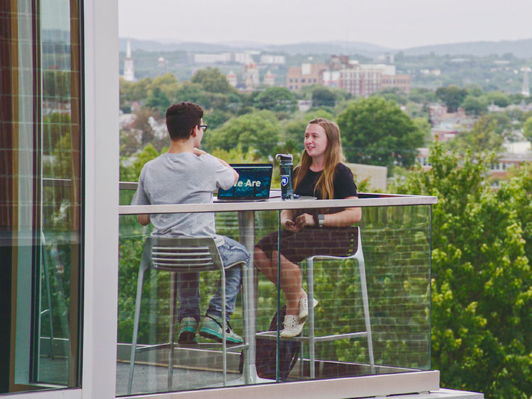 Male and female student sitting on balcony with York City skyline in the background.
