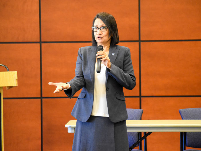 Ethnic female with glasses speaks in front of a group, not visible in photo.