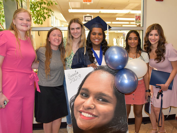 Six female students stading together, with one at the center wearing a graduation cap and gown.