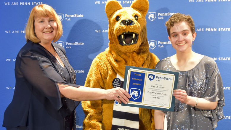 Famale presents an award to another a student, also female.  Person in Nittany Lion costume between the two.