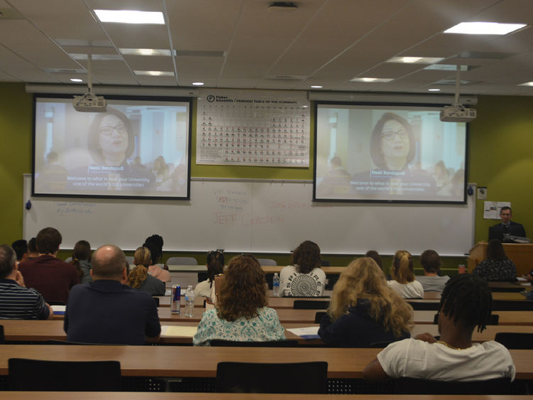 Photo from the back of a room with people seated and watching a screens during a program.