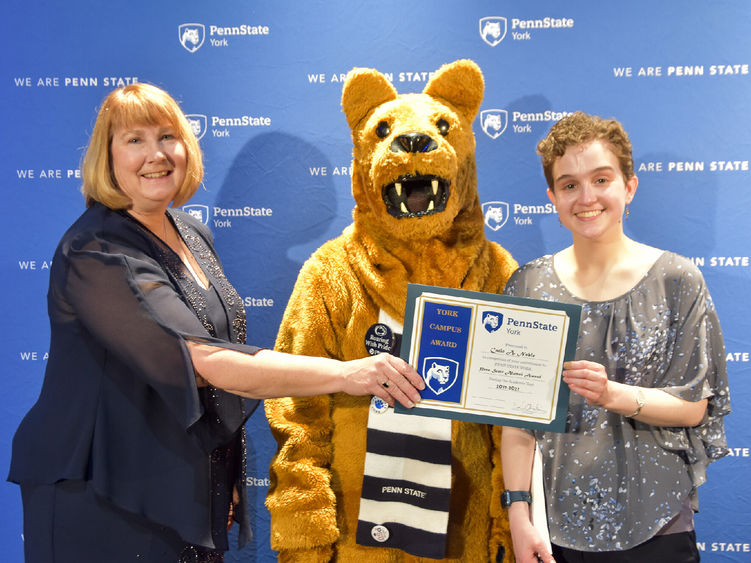 Famale presents an award to another a student, also female.  Person in Nittany Lion costume between the two.